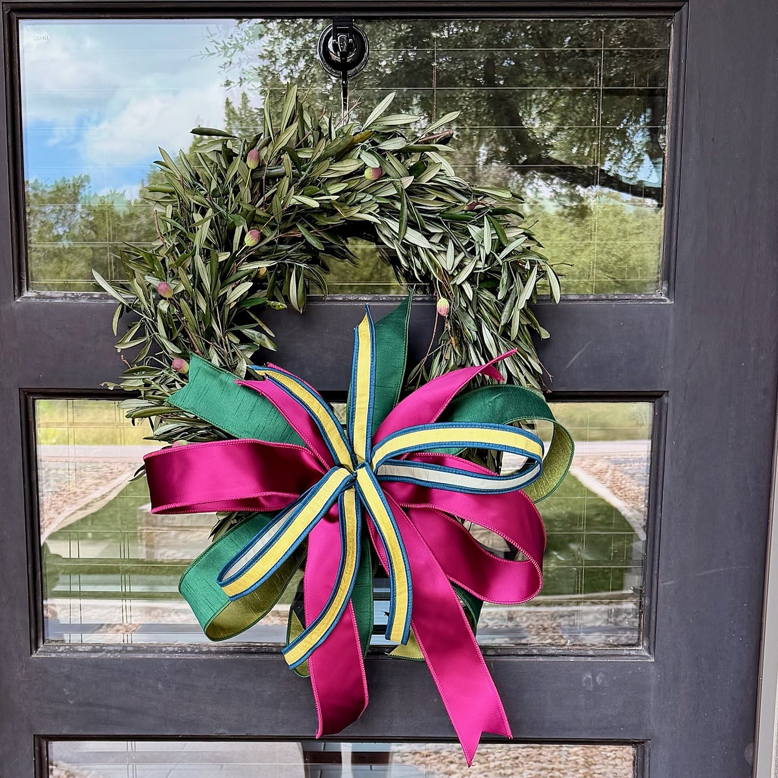Magenta, teal, and gold-striped ribbon bow tied onto a fresh olive wreath, hanging on a glass-paned front door reflecting trees and sky