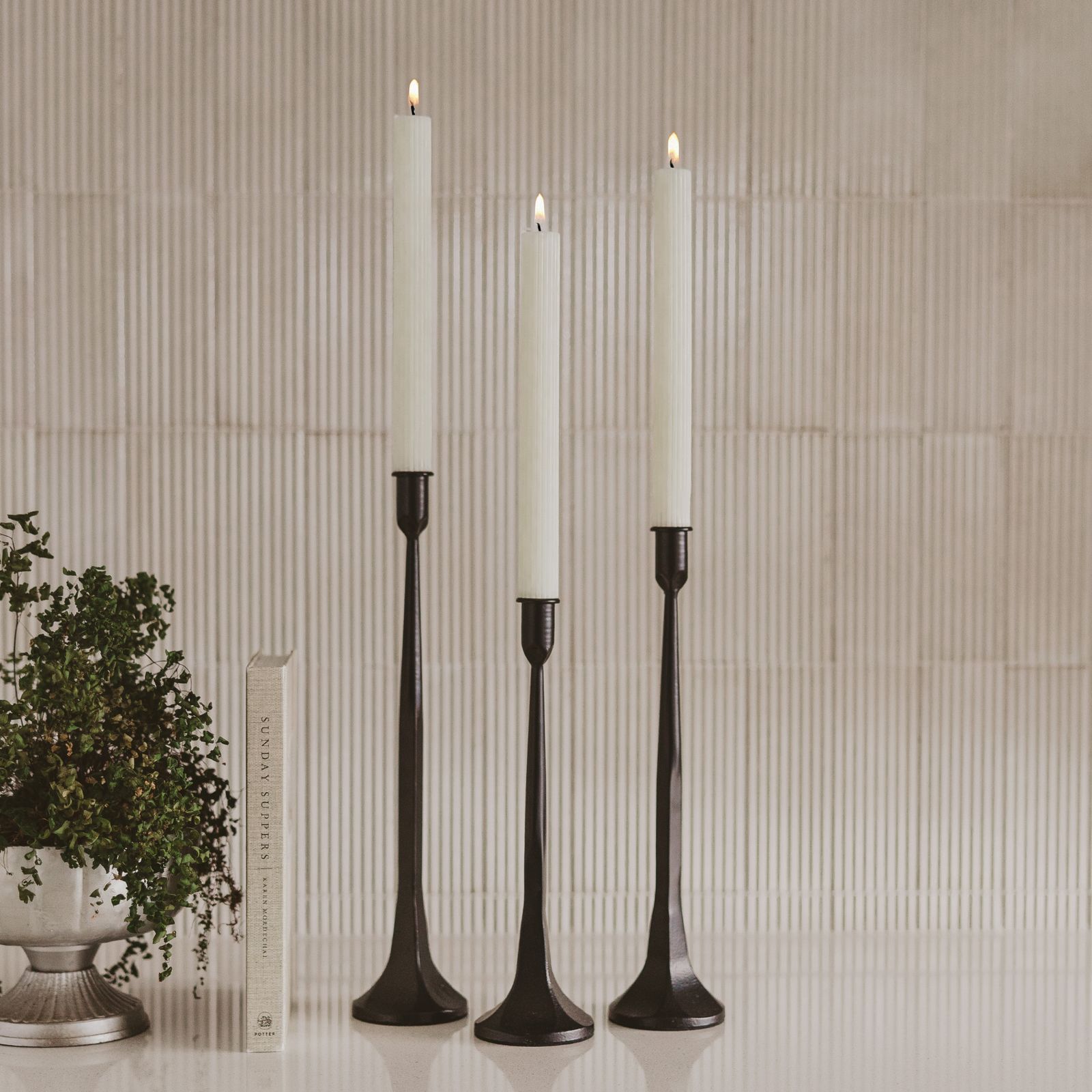 Set of three black cast iron taper candle holders in staggered heights, holding lit ivory candles against cream ribbed tile