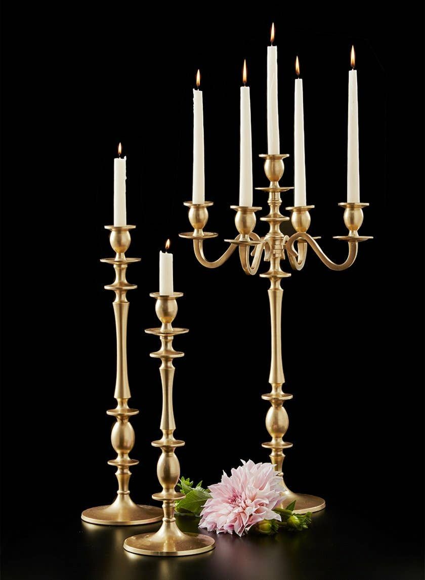 Tall gold candelabra with five lit taper candles beside two matching gold candlesticks and a single pink dahlia, on a black background