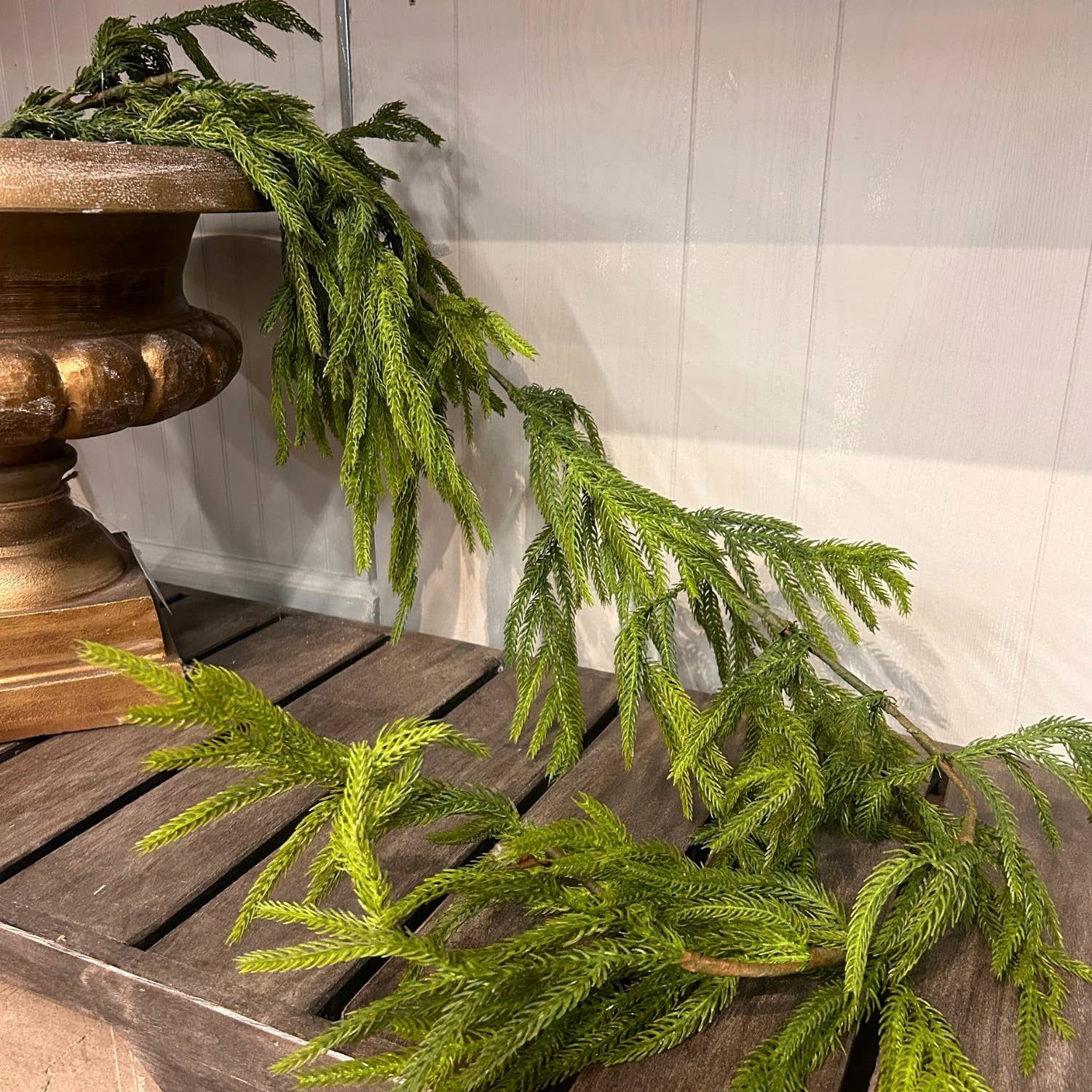 Norfolk pine garland draped from a gold urn onto a wooden slat bench